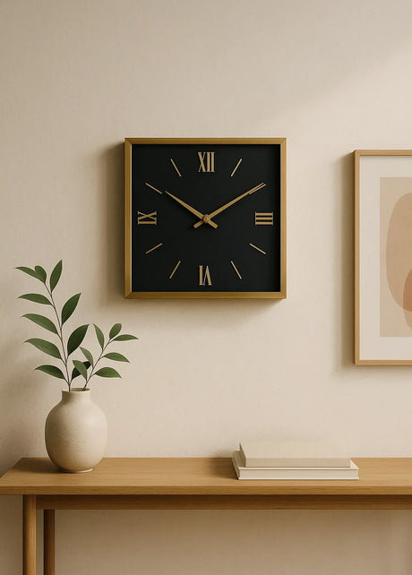 Square wall clock with black face, gold frame, and Roman numerals on a minimalist wall, above a console table with a vase and books.